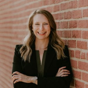 This is a headshot of Carly Wahl, Ph.D. The image shows a woman from the waist up, wearing a green shirt and black jacket. She is crossing her arms and standing next to a brick wall. She has blonde hair and is smiling for the camera. 